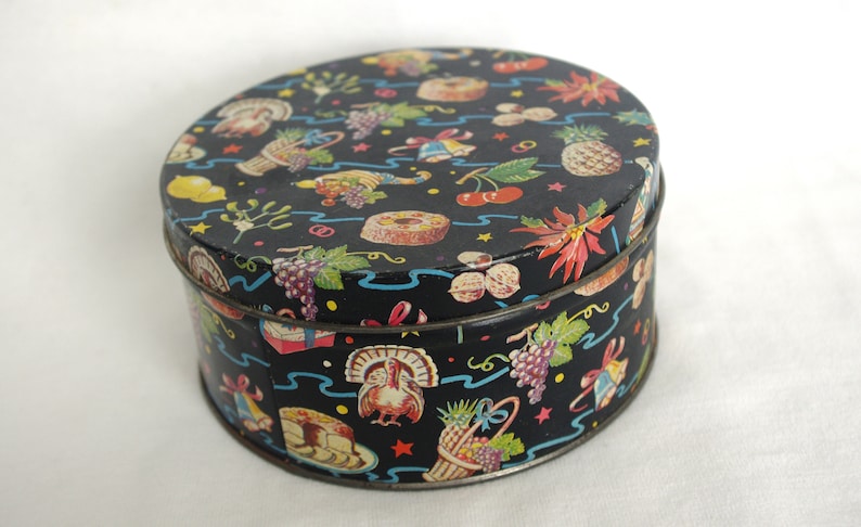 May include: A round, vintage tin with a black background and a colorful pattern of fruits, desserts, and festive decorations. The tin has a lid and a metal rim, perfect for storage or display.