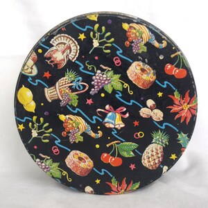 May include: Round, vintage tin with a black background and a colorful pattern of fruits, vegetables, and holiday symbols. The design includes a turkey, grapes, cherries, and a pineapple. The tin has a metallic rim.
