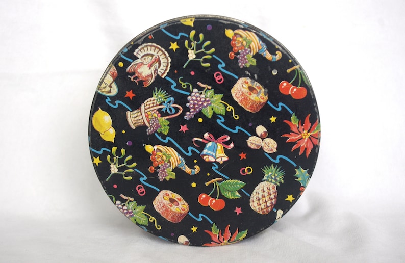 May include: Round, vintage tin with a black background and a colorful pattern of fruits, vegetables, and holiday symbols. The design includes a turkey, grapes, cherries, and a pineapple. The tin has a metallic rim.