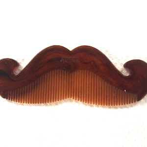 Puede incluir: Un peine marrón en forma de bigote con dientes finos. El peine tiene un diseño curvo en cada extremo, y los dientes son de un tono marrón más claro. El peine está hecho de un material liso y brillante.