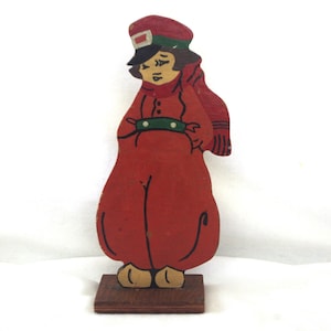 May include: A vintage wooden figurine of a person in a red coat, hat, and scarf. The figure has a painted face and is standing on a small wooden base. The figure's outfit is accented with green and black details.