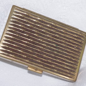 May include: A rectangular, gold-toned compact with a textured, ridged surface. The compact has a small clasp and appears to be made of metal. The design is simple and elegant, suitable for carrying small items.