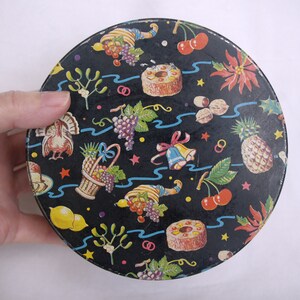 May include: A round, black container with a festive pattern. The design includes illustrations of fruits, nuts, and holiday symbols like bells and a turkey. The container is held by a hand.
