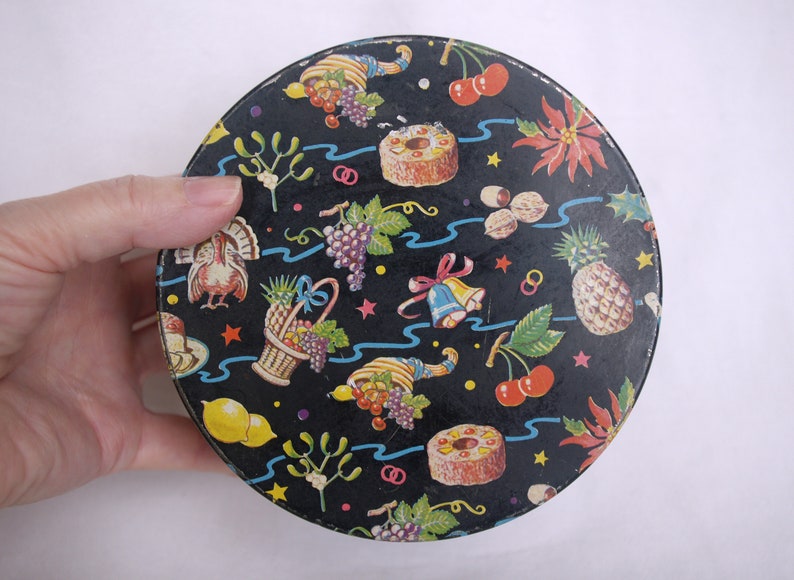 May include: A round, black container with a festive pattern. The design includes illustrations of fruits, nuts, and holiday symbols like bells and a turkey. The container is held by a hand.