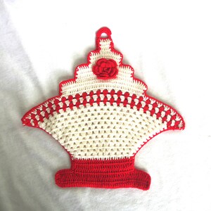 May include: A crochet potholder shaped like a basket with a red base and a cream-coloured body featuring a red checkered pattern. The top section is cream-coloured with a small red crocheted rose and a loop at the peak for hanging purposes.