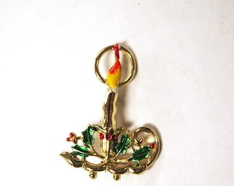 May include: A gold-coloured Christmas candle brooch with a yellow and red flame. The candle is decorated with green holly leaves and red berries. The brooch has a loop at the top for hanging.