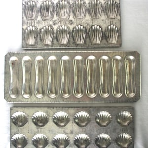 May include: Three vintage metal baking molds. The top and bottom molds feature shell-shaped cavities, while the middle mold has elongated, teardrop-shaped cavities. The molds are made of a silver-colored metal and are likely used for baking madeleines and other small pastries.