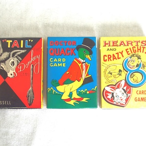 May include: Three vintage card game boxes featuring colorful illustrations. The first box is red and black with a donkey, titled Tail the Donkey Game by Russell. The second is blue with a duck in a top hat, titled Doctor Quack Card Game. The third is yellow with a monkey and rabbit, titled Hearts and Crazy Eights Card Games.