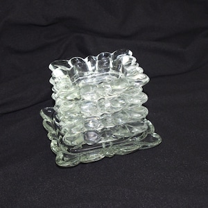 May include: A stack of clear glass coasters with a scalloped edge design. The coasters are square-shaped and have a textured surface. The stack is on a black background.