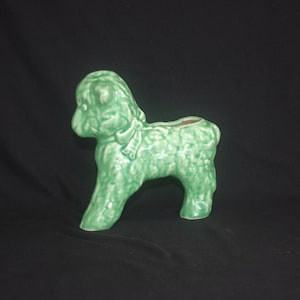Könnte beinhalten: Ein hellgrüner Keramik-Pudel-Pflanztopf. Der Pudel steht mit einer Schleife um den Hals. Der Pflanztopf hat eine strukturierte Oberfläche und eine kleine Öffnung auf dem Rücken für Pflanzen. Ein dekorativer Artikel.