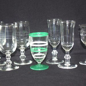 Può includere: Una collezione di calici in vetro trasparente, tra cui calici da vino e flûte da champagne, disposti su una superficie nera. Un bicchiere presenta strisce verdi e bianche. I bicchieri variano per forma e design.