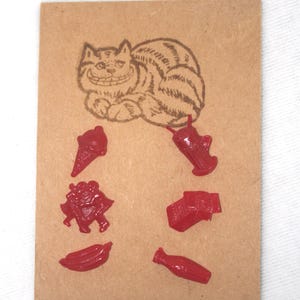 May include: A rectangular tan card featuring a brown ink illustration of a grinning cat at the top. Below the cat are six small, glossy red plastic charms shaped like an ice cream cone, a milkshake, two figures dancing, a gift, a banana, and a bottle.