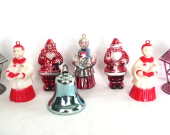 May include: A collection of vintage Christmas ornaments. The ornaments include red and white choir figures, Santa Claus figures, a silver bell, and two decorative lanterns. The ornaments are made of glass and metal and are arranged on a white surface.