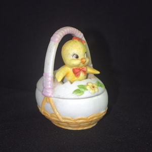 May include: A ceramic Easter basket with a yellow chick figurine inside. The basket is white with a woven texture at the base and a pink handle. The chick has a red bow and is emerging from a cracked egg.