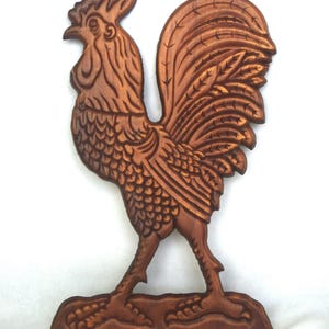 Può includere: Una placca da parete in legno intagliato, piatta e di colore marrone, a forma di gallo. Il gallo è raffigurato di profilo, rivolto verso sinistra, con dettagli intagliati sulle piume del collo, del corpo e della coda, su una base.