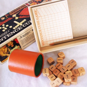 May include: Vintage Scrabble Sentence Cube Game box set. The box is open, revealing a score pad, pencil, orange cup, and wooden word cubes. The box has the words "SCRABBLE SENTENCE CUBE GAME" printed on it.
