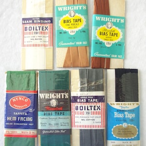 Könnte beinhalten: Eine Sammlung von Vintage-Nähzubehör, darunter Schrägband, Nahtband und Saumaufschlag. Die Verpackungen zeigen verschiedene Farben wie Beige, Braun, Grün und Schwarz, mit Textetiketten, die die Produktart und Marken wie "Wright's" angeben.