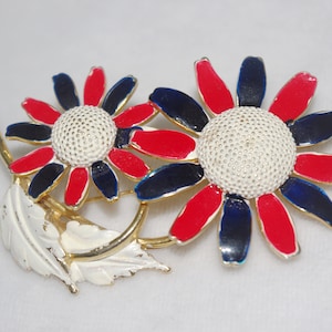 May include: A vintage floral brooch featuring two daisy-like flowers with red, navy, and white petals. The brooch has a gold-tone stem and a white leaf, creating a patriotic color scheme.