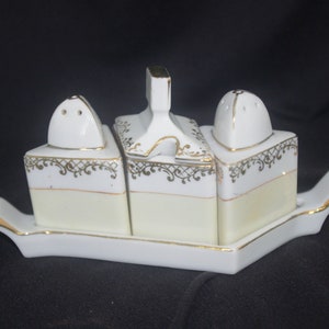 Puede incluir: Un juego de salero y pimentero de porcelana blanca con un azucarero a juego, todo sobre una bandeja rectangular. El juego presenta una base color crema, filigrana dorada y ribetes dorados.