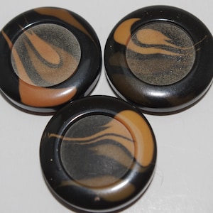 May include: Three round, black and tan decorative objects with a swirling pattern. Each has a central, lighter-colored circle. The objects are arranged in a triangular pattern against a white background.