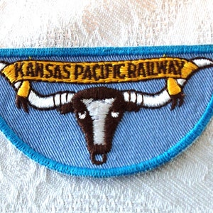 May include: Embroidered patch with a light blue background and a turquoise border. The patch features a brown and white longhorn skull with the text "KANSAS PACIFIC RAILWAY" in gold lettering on a yellow banner.