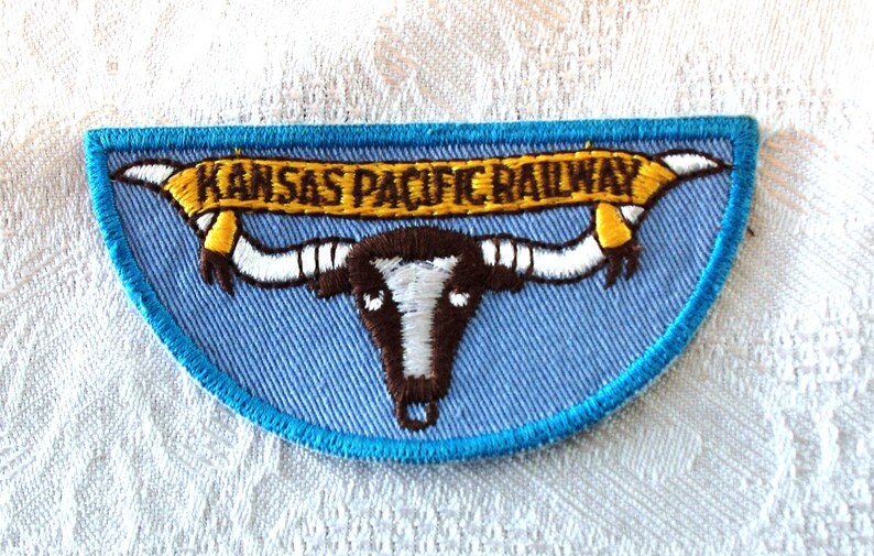 May include: Embroidered patch with a light blue background and a turquoise border. The patch features a brown and white longhorn skull with the text "KANSAS PACIFIC RAILWAY" in gold lettering on a yellow banner.