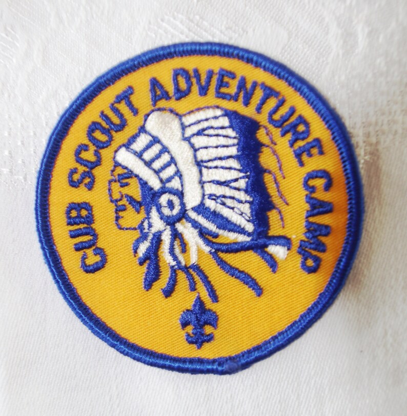 May include: A circular patch with the text "CUB SCOUT ADVENTURE CAMP" in blue lettering. The patch features a Native American chief in profile, wearing a headdress. The patch is yellow with a blue border and a blue fleur-de-lis.
