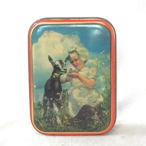 May include: A rectangular metal tin featuring a printed illustration of a child with blonde hair wearing a white dress, kneeling in a field of yellow flowers while feeding a small black and white goat. The tin has a bright red and gold border.