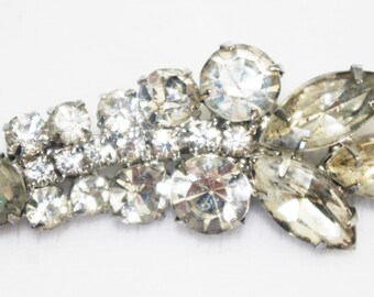 May include: A silver-toned brooch featuring a floral design with clear and smoky-colored rhinestones. The brooch has a mix of round and marquise-shaped stones, creating a sparkling, elegant accessory.
