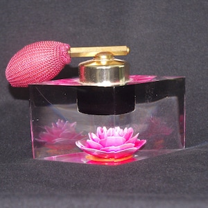 Può includere: Un flacone di profumo rettangolare trasparente con un tappo color oro e un vaporizzatore rosa. La bottiglia presenta un disegno floreale rosa all'interno. Lo sfondo è scuro.
