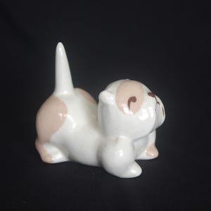 May include: A small ceramic figurine of a cat in a playful pose with its tail pointed upwards. The cat is primarily white with light tan patches on its back and head, featuring simple brown painted eyes and a nose on its smooth, glossy surface.