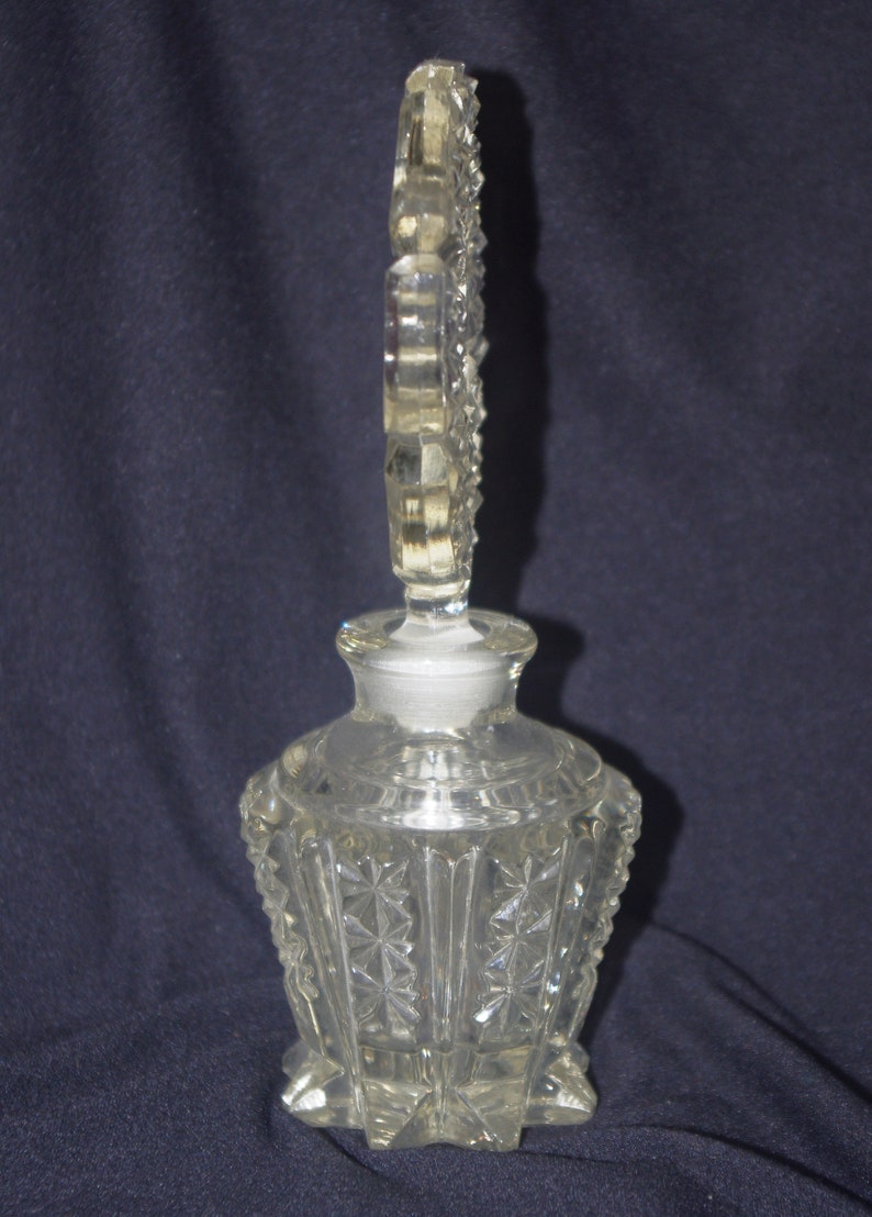 Pu&ograve; includere: Flacone di profumo in vetro trasparente con tappo decorativo. La bottiglia ha un design a stella sfaccettato sul corpo e un tappo alto e ornato. La bottiglia &egrave; probabilmente vintage e aggiunge un tocco di eleganza.