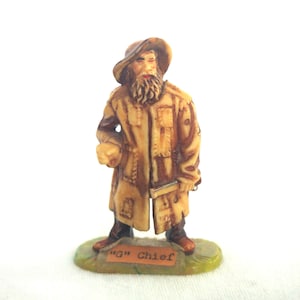 May include: A small figurine of a man in a long coat and hat, with a long beard. The figurine is tan and brown, with a green base. The text "G" Chief is printed on the base.
