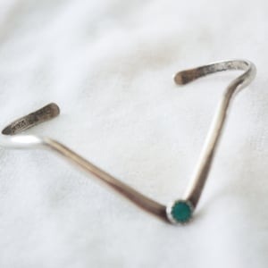 Puede incluir: Un brazalete plateado en forma de V con una pequeña piedra turquesa redonda en el centro. Los extremos del brazalete se curvan hacia arriba. La palabra "STERLING" está estampada en un extremo. El brazalete está sobre una superficie blanca.