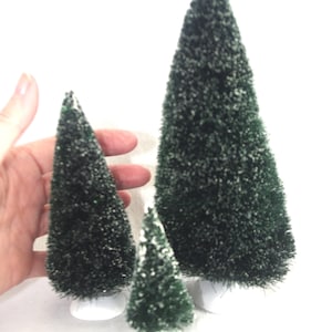 Può includere: Tre alberi di Natale in miniatura a spazzola per bottiglie di diverse dimensioni. Gli alberi sono verde scuro con una spolverata di neve bianca e poggiano su basi bianche. Gli alberi sono disposti in fila su uno sfondo bianco.