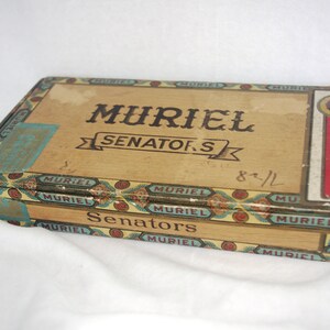 May include: A vintage wooden cigar box with the text "MURIEL SENATORS" in black lettering. The box has decorative trim with red, blue, and gold accents. The box is rectangular and appears to be aged.