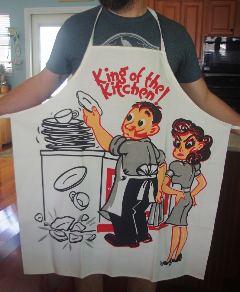 May include: White apron with a cartoon illustration of a man and woman in a kitchen setting. The apron features the text "King of the Kitchen!" in red. The man is depicted dropping dishes, while the woman looks displeased.