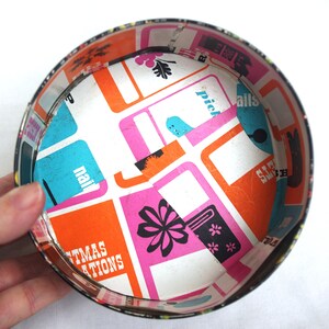 May include: A vintage, round metal container with a colorful interior. The inside features a white background with orange, pink, and blue rectangular shapes. The container's rim is black, and the interior design includes the text "Christmas Creations."