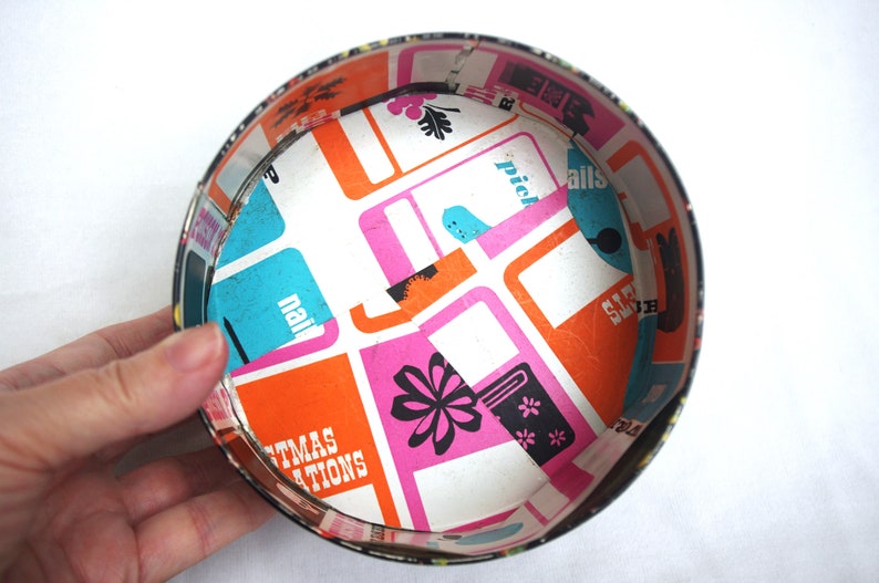 May include: A vintage, round metal container with a colorful interior. The inside features a white background with orange, pink, and blue rectangular shapes. The container's rim is black, and the interior design includes the text "Christmas Creations."