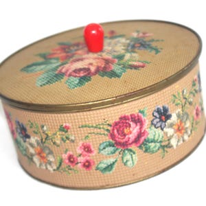 May include: Oval-shaped vintage box with a tan fabric exterior and floral needlepoint design. The lid features a cluster of roses and other flowers. The sides are adorned with a continuous floral pattern. A red knob serves as the handle.