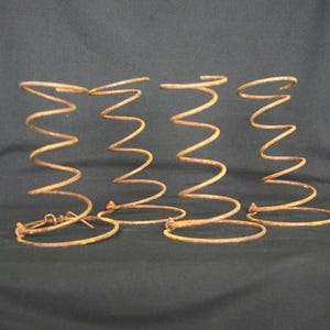 May include: Four rusty metal springs with a zigzag design. The springs are a warm, aged brown color and stand upright against a dark background. These springs could be used for furniture or other industrial applications.