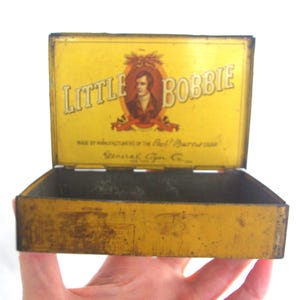 May include: Vintage yellow metal box with the words "LITTLE BOBBIE" in white lettering. The box features an illustration of a man in a brown oval frame. The box is open, revealing an empty interior. The box is held by a hand.