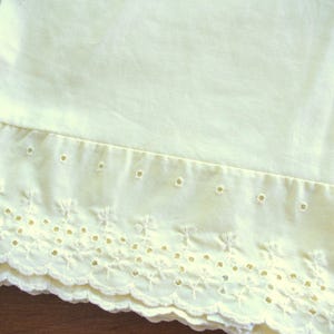 Può includere: Primo piano di un tessuto giallo pallido con un bordo smerlato e traforato decorativo. Il tessuto sembra essere un cotone o un lino leggero, con una consistenza morbida e leggermente stropicciata. I dettagli traforati aggiungono un tocco delicato e vintage al tessuto.