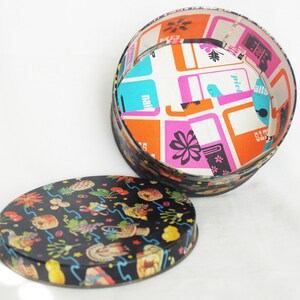 May include: A vintage round tin box with a lid. The lid is black with colorful illustrations. The interior of the box is white with a colorful geometric pattern. The box is open, revealing the interior design.