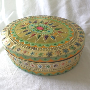 May include: An oval metal storage tin featuring a gold-toned lid with a central pink jewel accent surrounded by colorful starburst patterns. The sides and lid are decorated with scalloped borders, green polka dots, and small floral motifs.