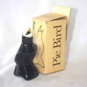 May include: A glossy black ceramic pie bird with a white beak, positioned next to its original light yellow cardboard box. The box features a line drawing of the bird and the text "Pie Bird" printed in dark brown ink on the side panel.