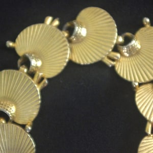 Pode incluir: Colar dourado com pingentes em forma de leque com ranhuras radiais. Cada pingente é conectado por pequenas argolas texturizadas. O colar tem uma estética vintage.