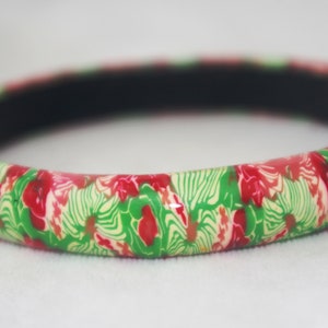 May include: A round bangle bracelet with a floral pattern in red, green, and cream colors. The bracelet has a black interior and is set against a white background. This accessory is suitable for everyday wear.