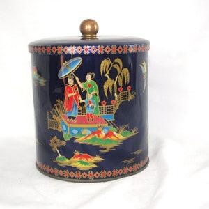 May include: A vintage, cylindrical tin canister with a dark blue background and an Asian-inspired design. The lid is dark blue with a gold knob. The canister features a colourful scene with figures, trees, and bridges.