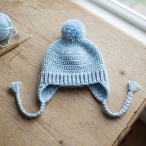 May include: A light blue crocheted hat featuring a ribbed brim, ear flaps with braided tassels, and a matching pom-pom on top. The hat is constructed from yarn and displays a consistent stitch pattern throughout the main body and ear flaps.